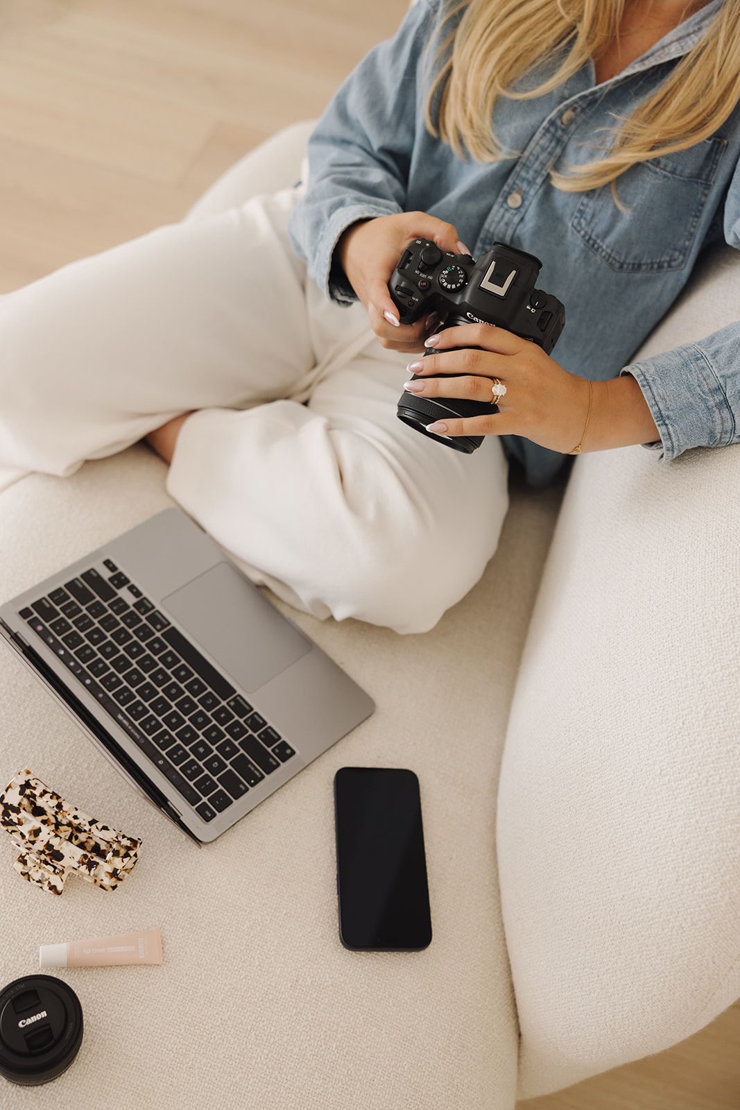 Woman in denim shirt holding Canon camera, sitting with laptop and smartphone on white chair.