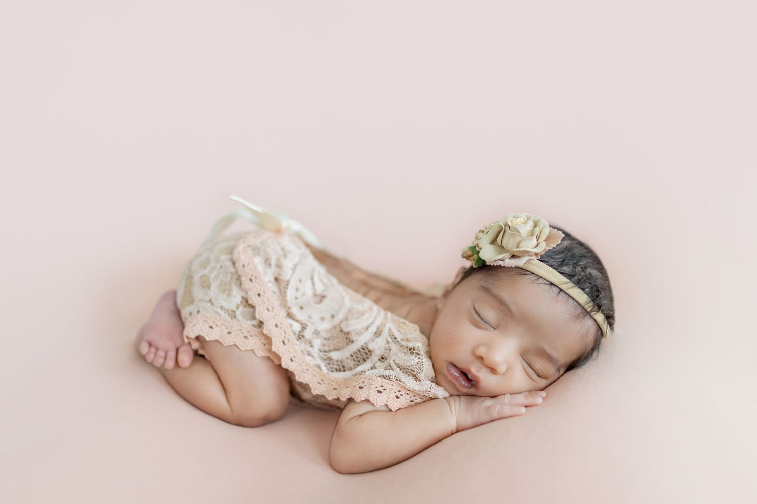 Sleeping newborn baby girl in lace romper and floral headband posed on pink backdrop.