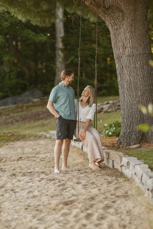 A couple shares a romantic moment outdoors, woman sitting on a tree swing, man standing beside her on sandy path.
