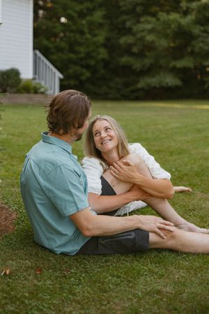 A couple sits together on green grass, smiling and embracing near a white house surrounded by lush trees.