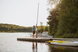 Couple standing on a wooden dock by a sailboat on a calm lake surrounded by lush green trees at sunset.