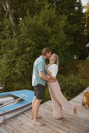 Couple sharing a romantic kiss on a wooden dock by a blue boat, surrounded by lush green trees at sunset.