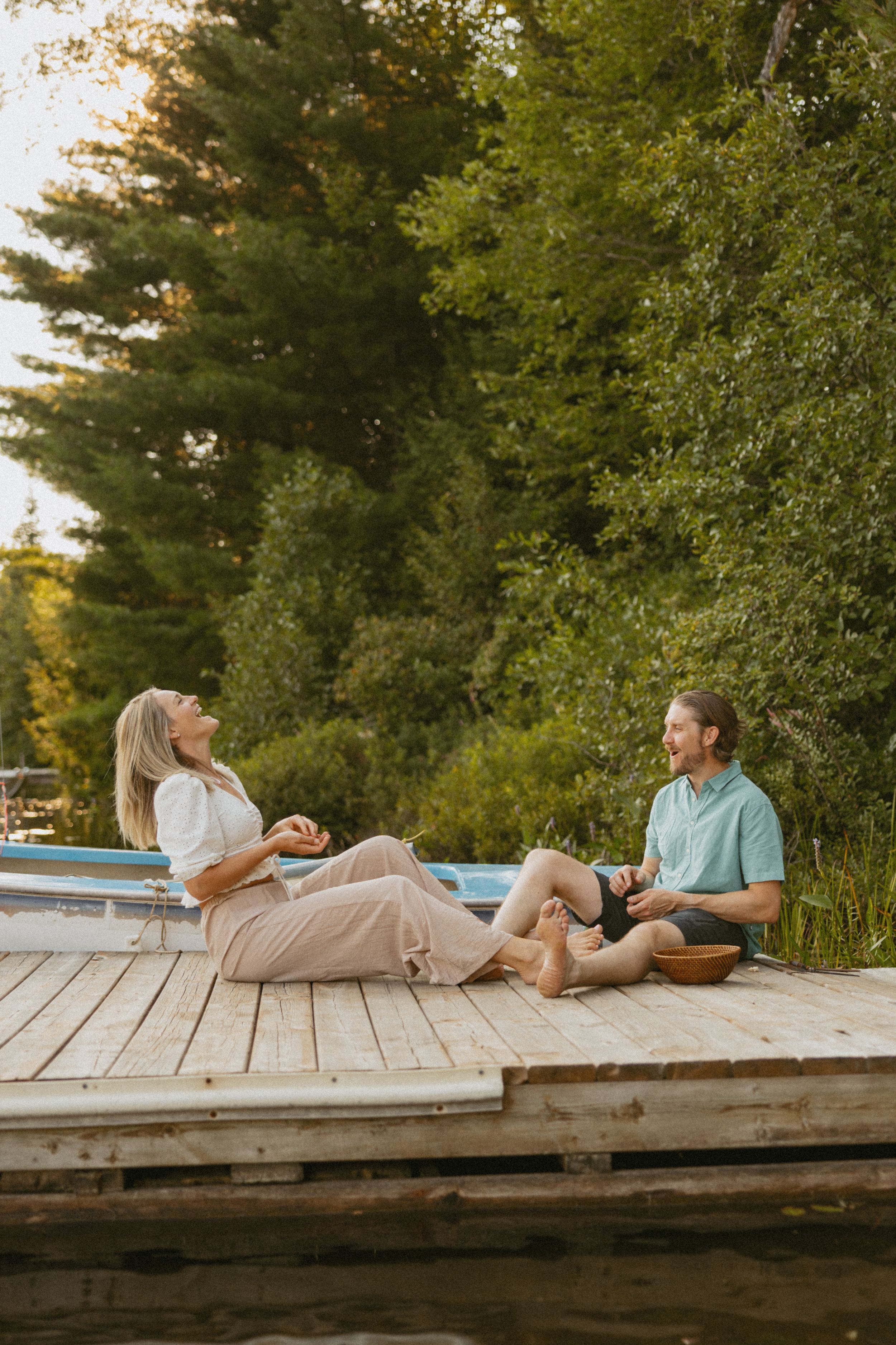 Couple sitting on a wooden dock by a lake, laughing and relaxing near a boat surrounded by lush green trees.