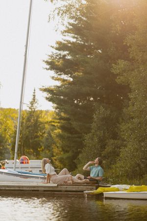 Couple relaxing on a wooden dock by a sailboat at golden hour, surrounded by lush green trees near a calm lake.