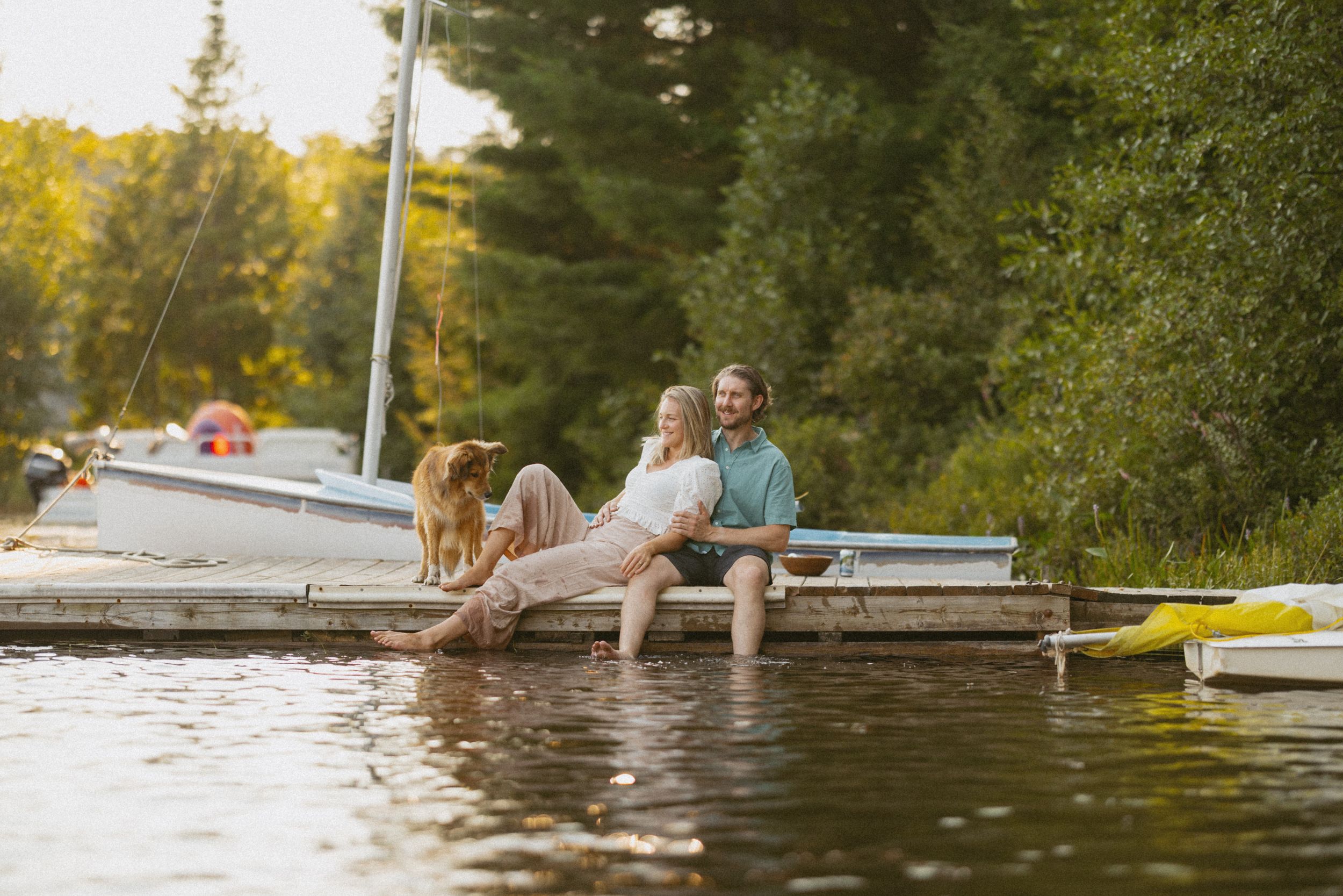 Couple sitting on a lakeside dock with their dog, sailboat nearby, enjoying a sunny summer day surrounded by lush green trees.