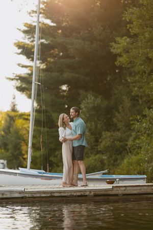 Couple embracing on a dock beside a sailboat, bathed in golden sunset light with green trees in the background.