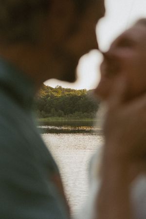 Romantic couple nearly kissing with a serene lake and lush green trees reflected in the water behind them.