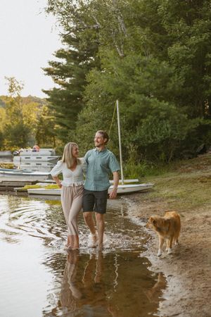 Couple walks barefoot along a lakeside shore with their golden retriever dog near docked boats and lush green trees.