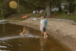 Couple standing on sandy lakeside beach while golden retriever splashes in shallow water near wooded property.