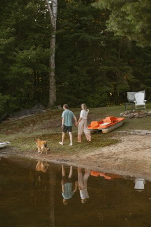 Couple holding hands with dog by lakeside orange pedal boat, surrounded by lush green forest at dusk.