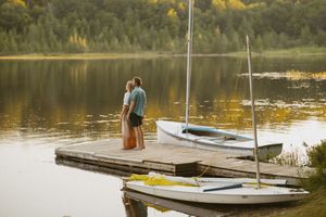 Couple embracing on a wooden dock by a calm lake at sunset, with sailboats and autumn foliage in the background.