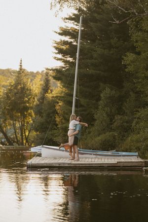 Couple embracing on a wooden dock beside a sailboat, surrounded by lush green trees reflecting on a calm lake at sunset.