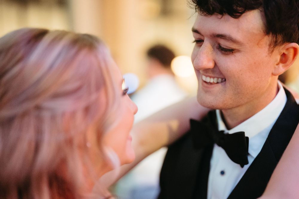 A smiling groom in a black tuxedo gazes at a blonde woman during an outdoor wedding celebration.