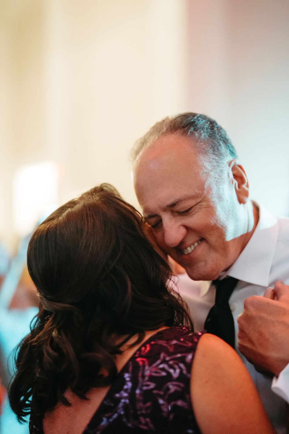 An older man smiles warmly while sharing a tender dance with a woman in a purple dress at a formal event.