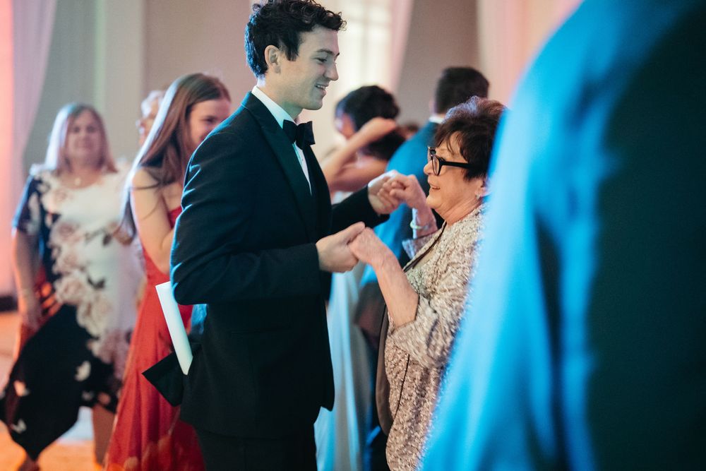 A young man in a tuxedo dances with an older woman in a floral dress at a formal event.