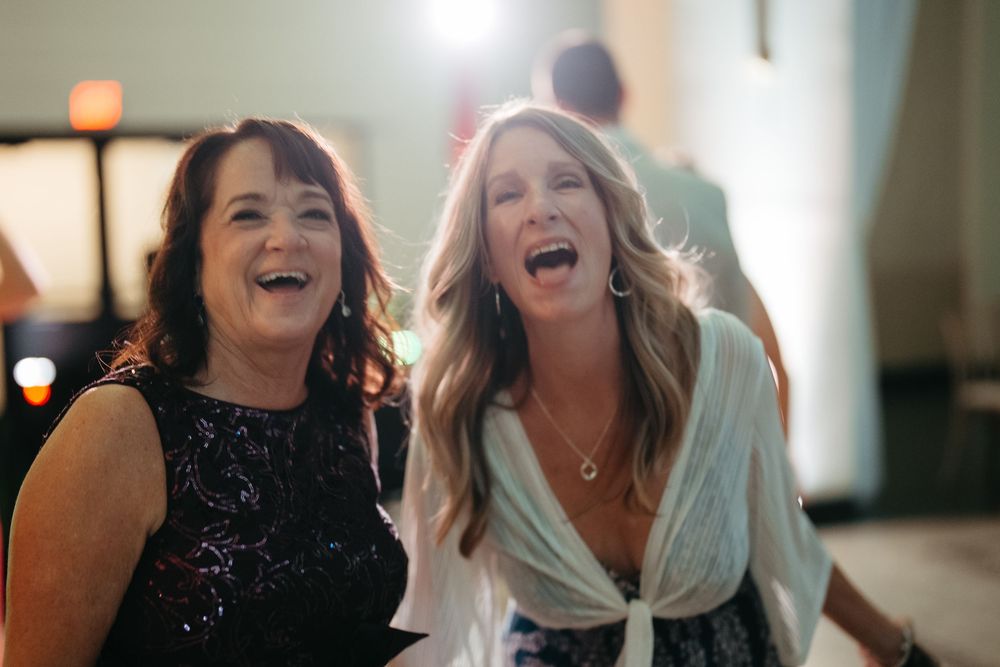Two women laughing and singing joyfully together at a festive indoor event, one in black sequins, one in white wrap top.