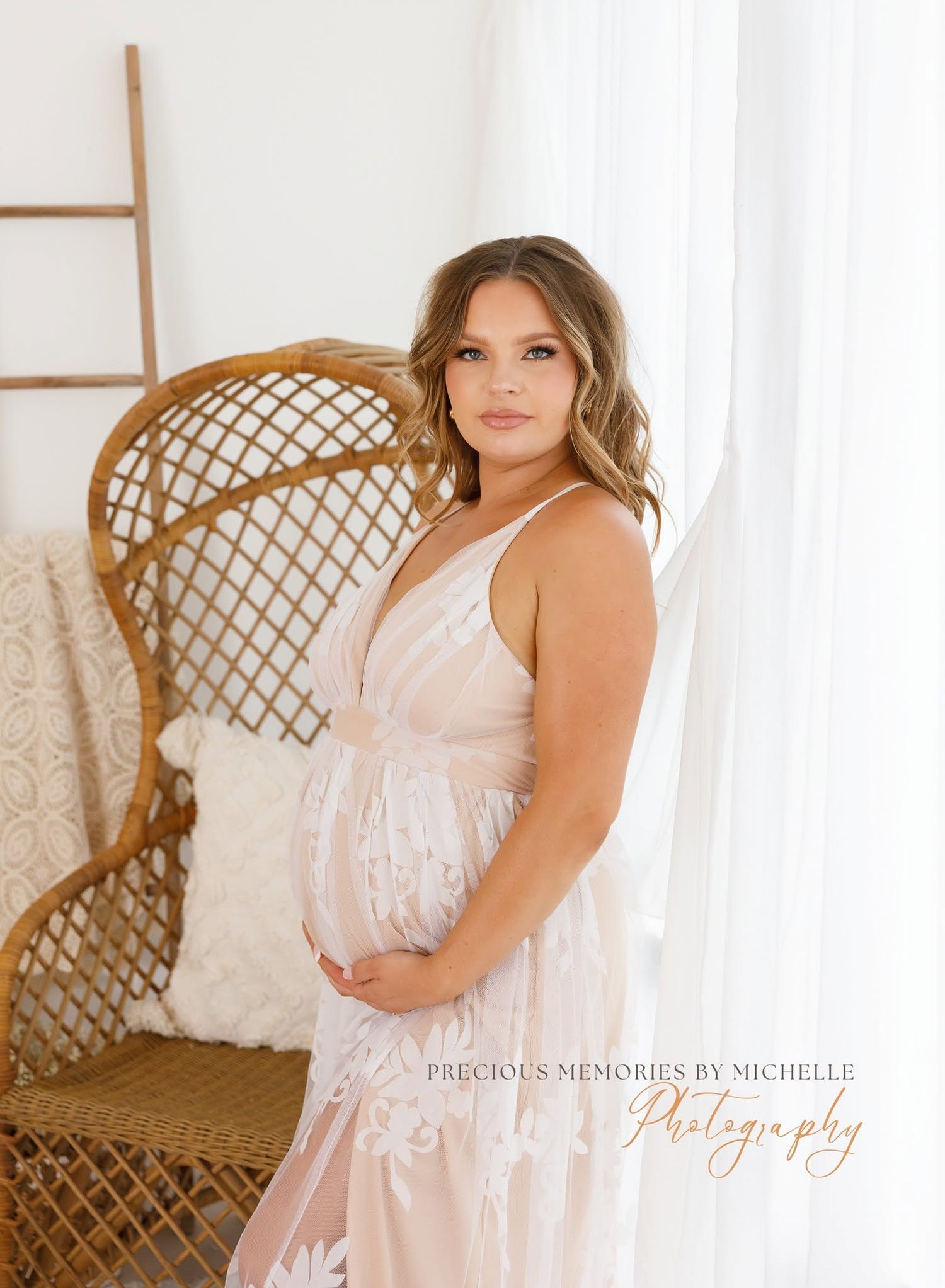 Pregnant woman in white lace gown poses elegantly beside a wicker chair near sheer curtains in a bright studio.