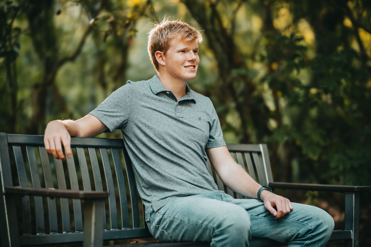Young man in gray polo shirt sitting relaxed on wooden bench in lush green park setting.