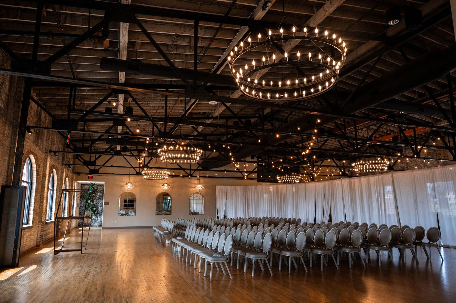 Elegant wedding venue with circular chandeliers, draped white curtains, and rows of chairs on hardwood floors.