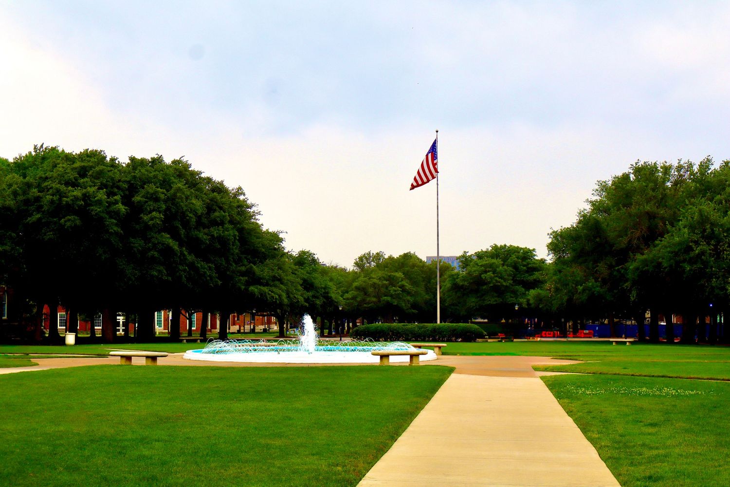 American flag flying over a scenic park with a fountain, green lawn, and tree-lined path on a cloudy day.