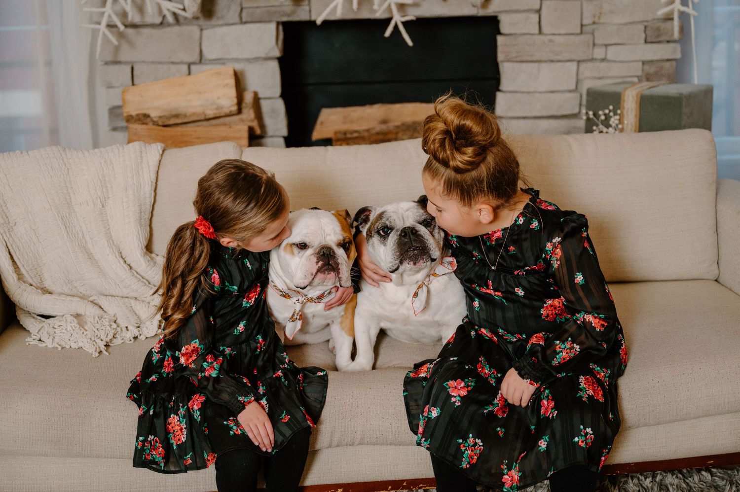 Two girls in matching floral dresses sit on a couch with two English Bulldogs near a cozy fireplace.