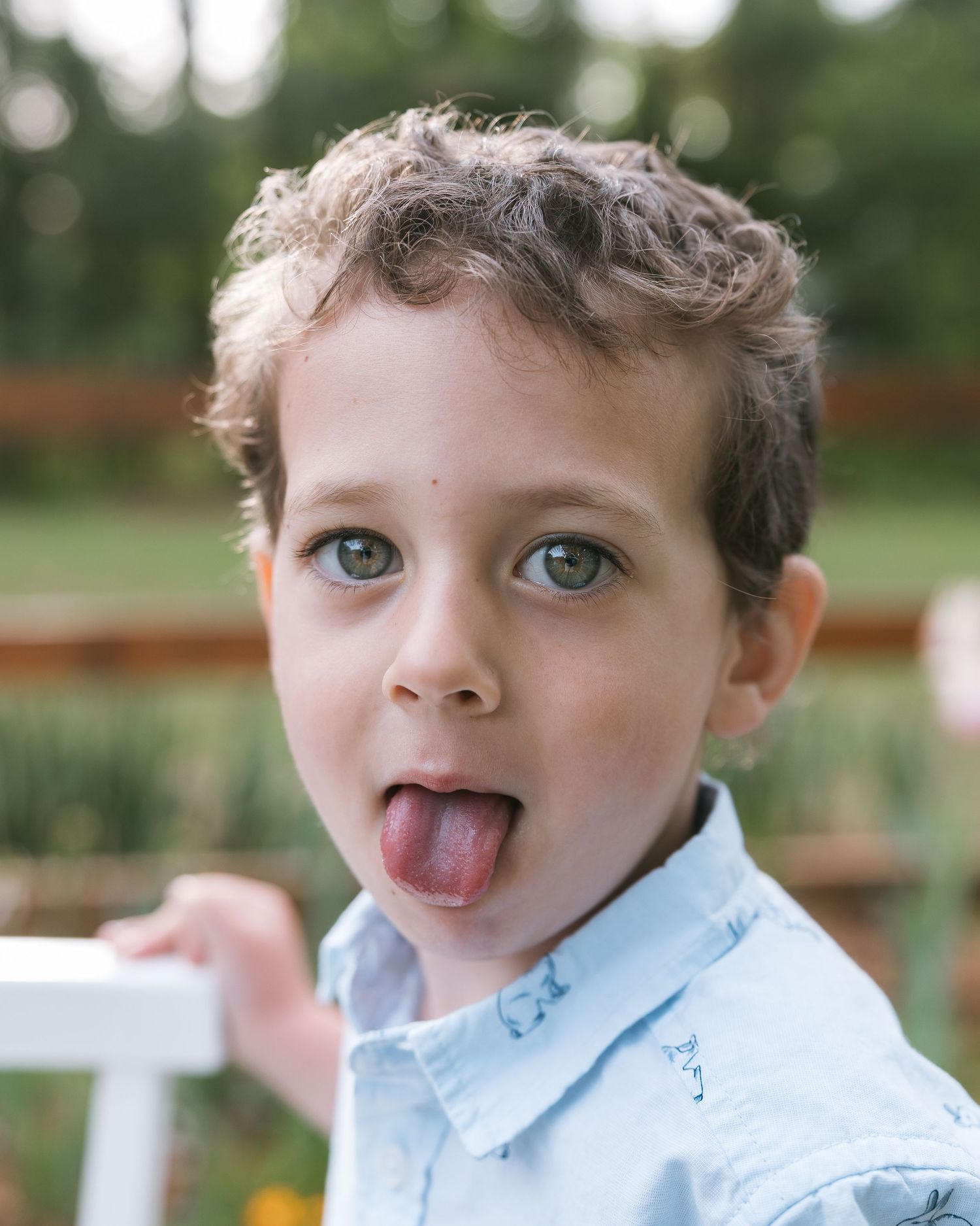 Young boy sticking his tongue out playfully while wearing a light blue shirt outdoors.