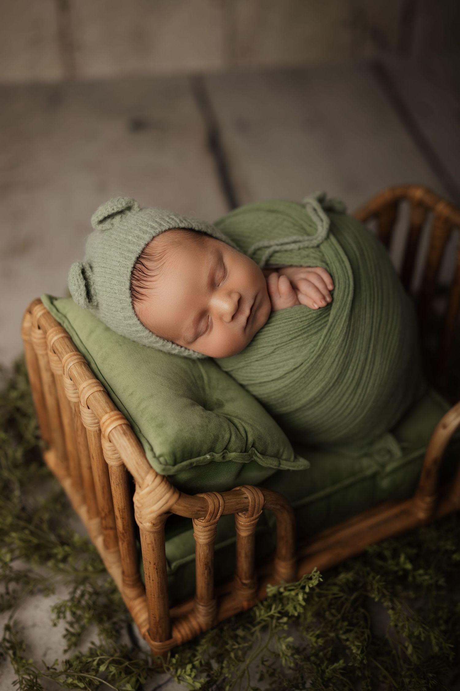 Newborn baby wrapped in green swaddle and bonnet sleeping peacefully in a small wicker bed with green cushion.