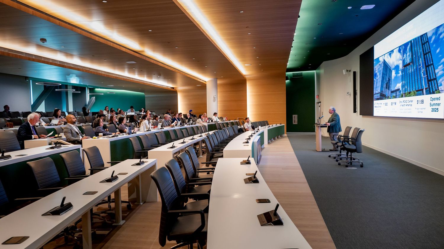 Modern lecture hall with tiered seating, wood paneling, and a large presentation screen during a professional conference.