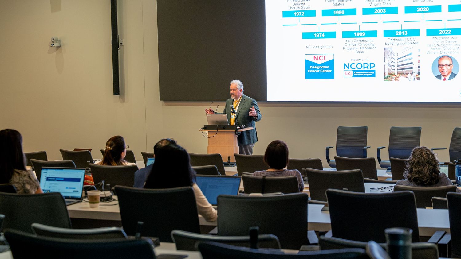 Speaker presenting at a corporate conference in a modern lecture hall with attendees and a colorful data slide projected behind.