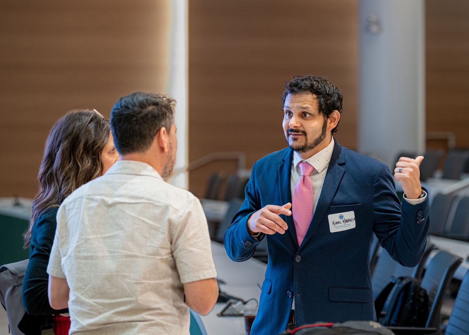 Professional man in blue suit and pink tie gestures while networking with colleagues at a business conference.