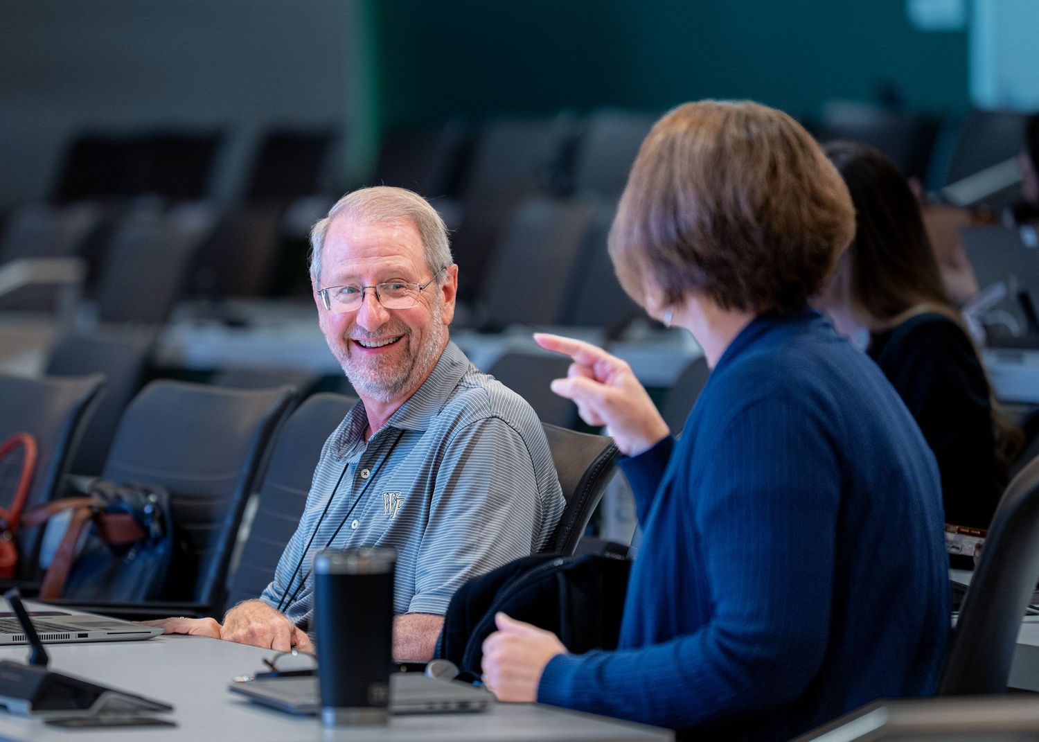 Two colleagues smile and chat at a conference table in a modern lecture hall setting.