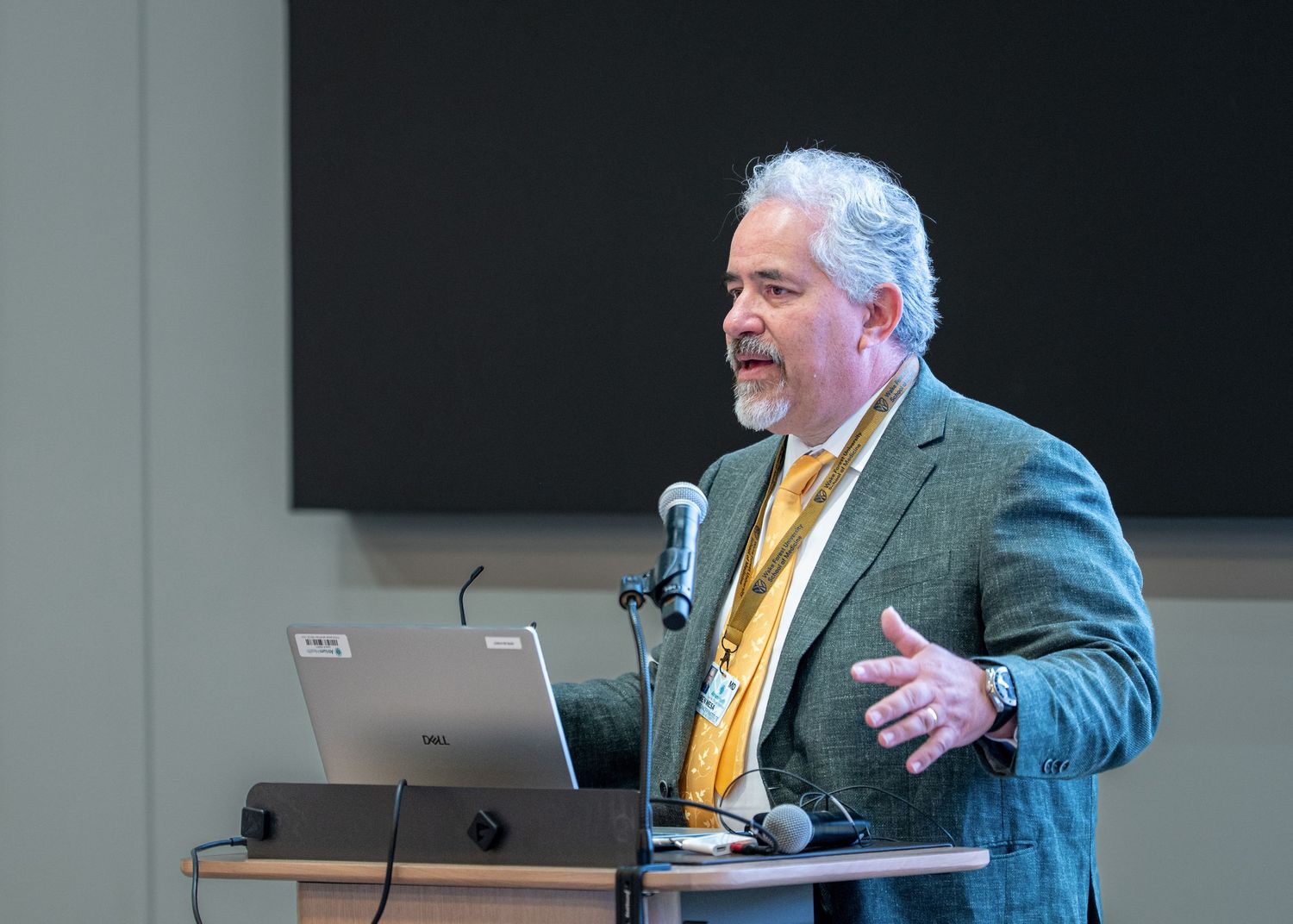 Professional speaker presenting at podium with laptop, wearing gray blazer and yellow lanyard in conference room.