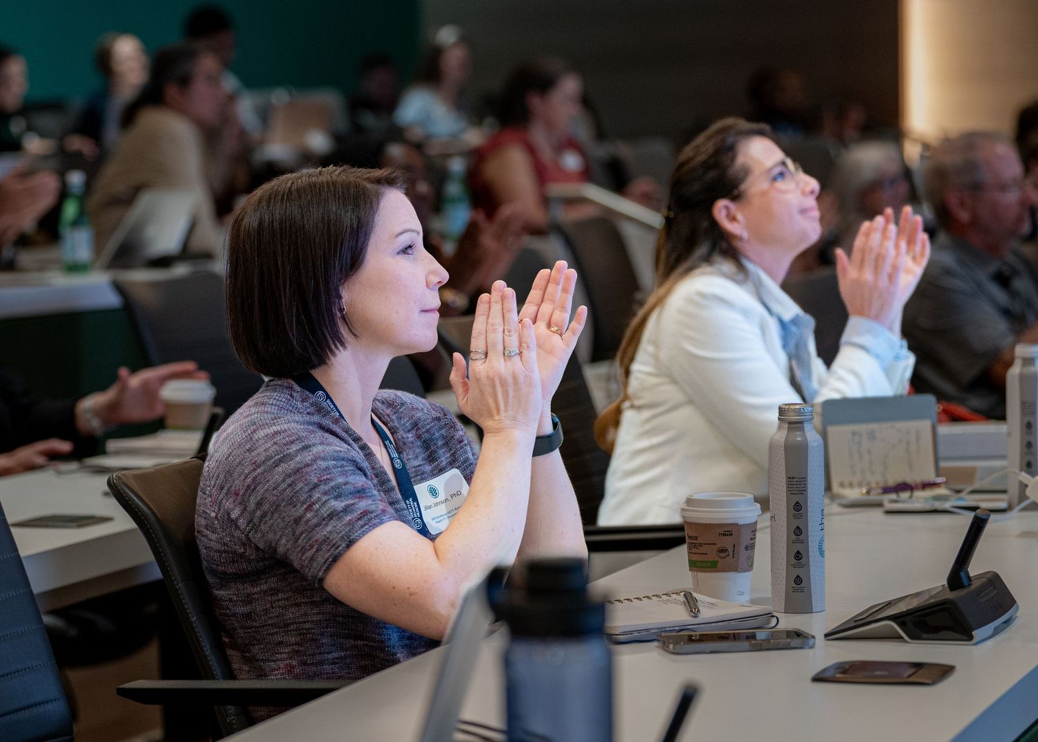 Audience members applaud during a professional conference session, seated at tables in a modern meeting room.