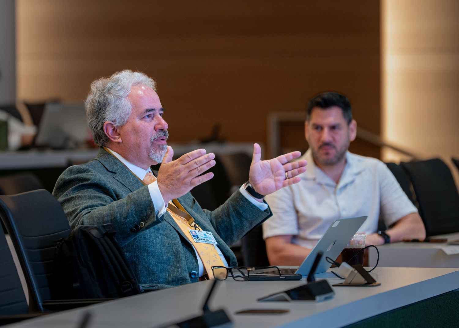 Man in suit gestures expressively during business meeting while colleague listens attentively in modern conference room.