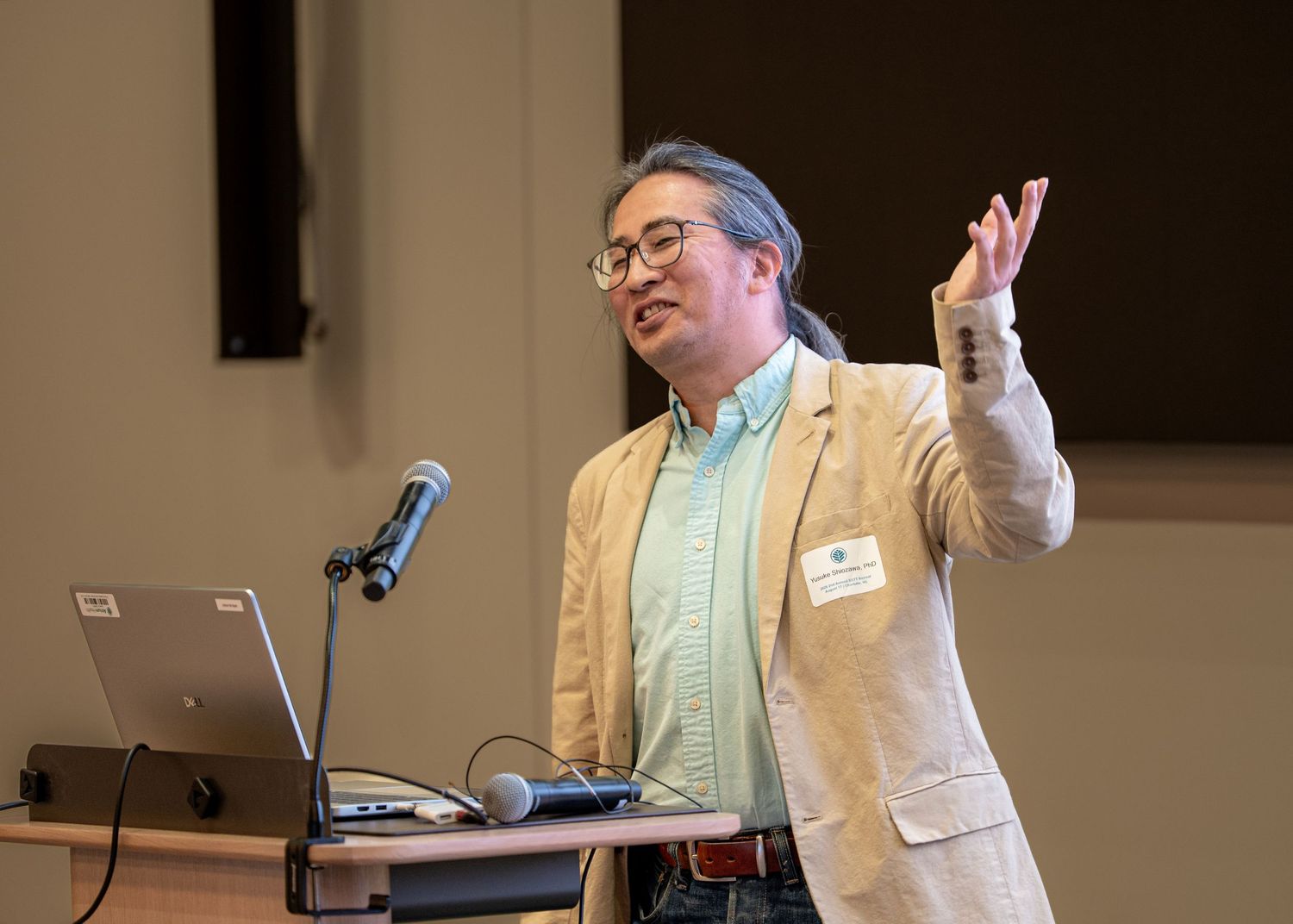 Man in beige blazer gesturing while presenting at a conference podium with a laptop and microphone.