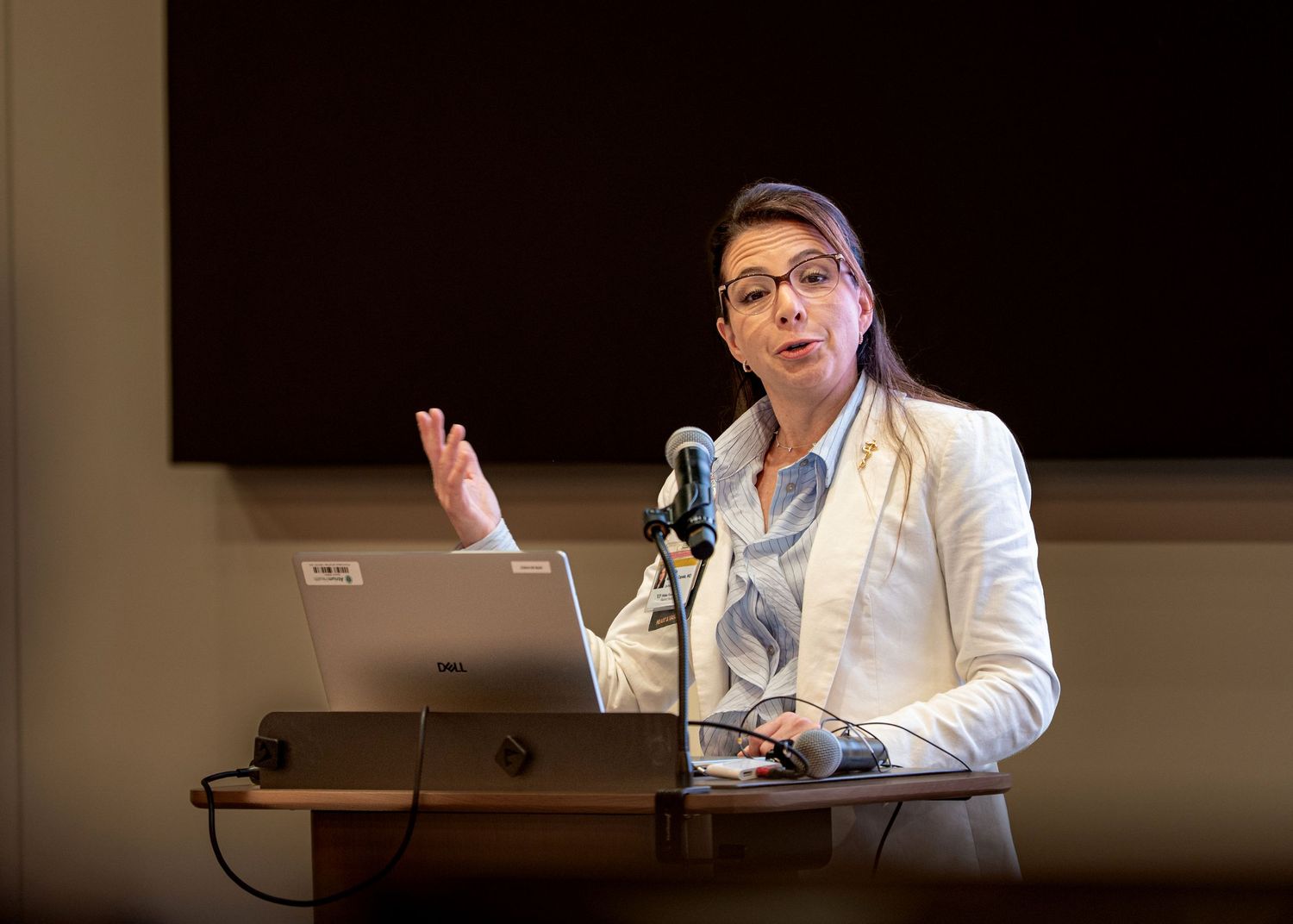 Woman in white blazer presenting at podium with laptop and microphone in professional conference setting.