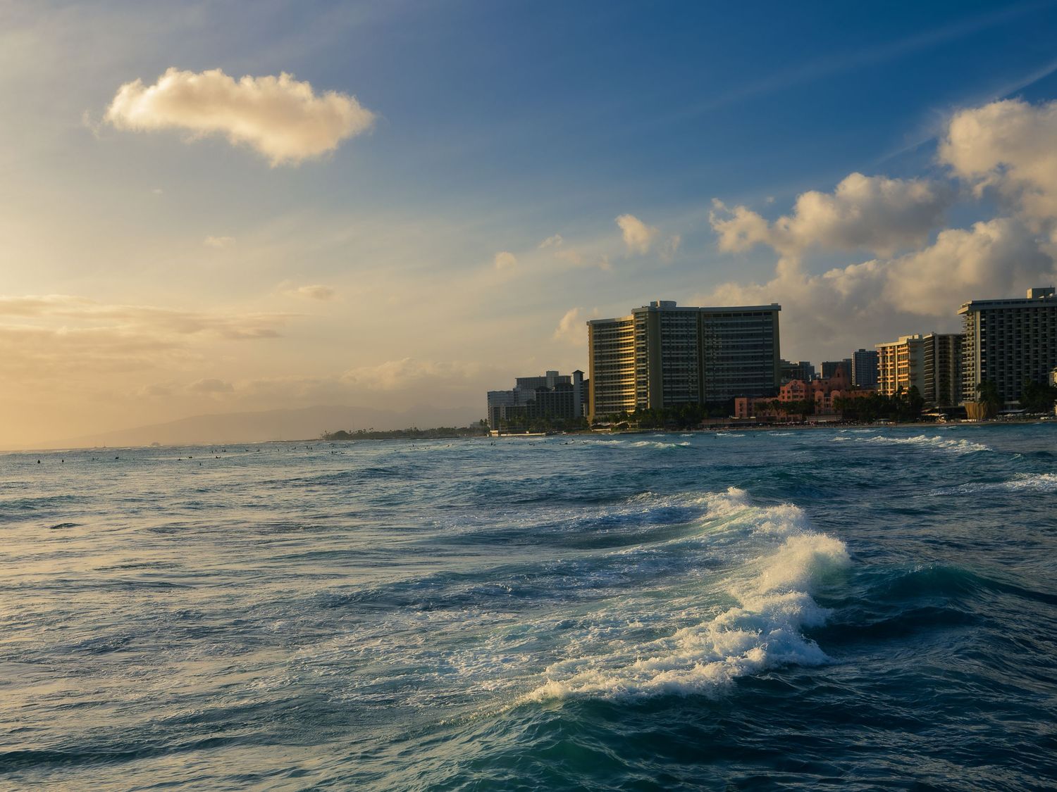 Waves roll toward the Waikiki Beach shoreline in Honolulu at golden hour, with beachfront hotel towers and distant surfers along the coast.