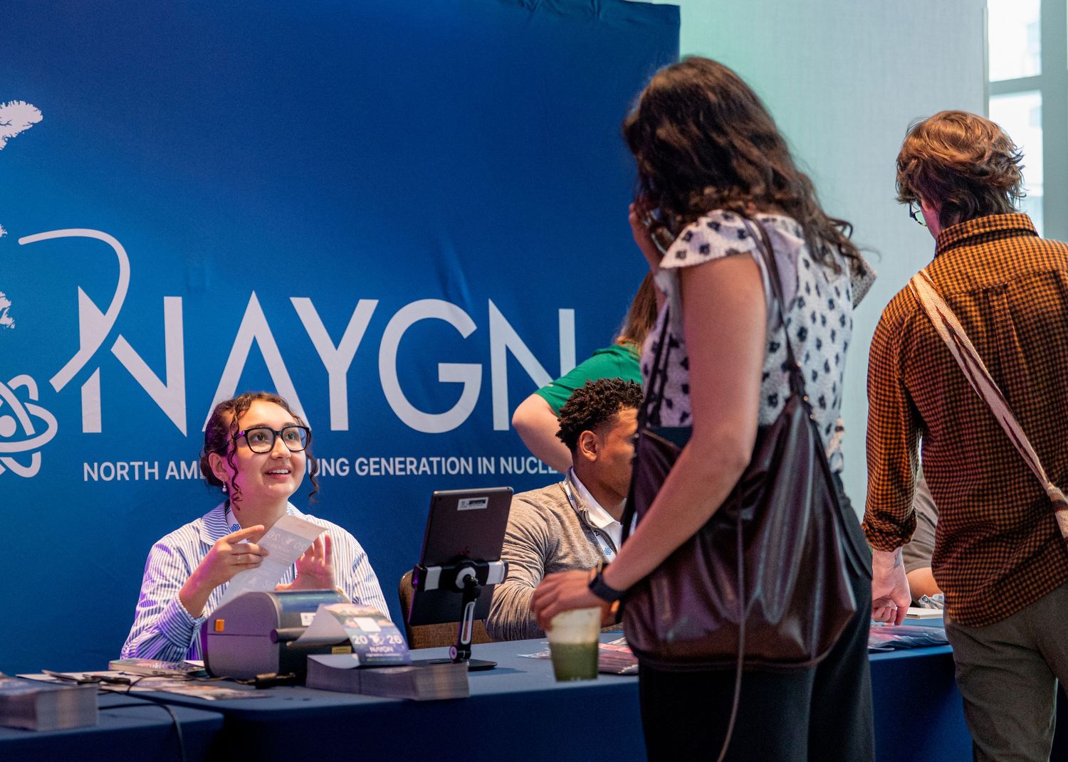 NAYGN booth at a nuclear energy conference with a smiling representative assisting attendees at a blue branded table.