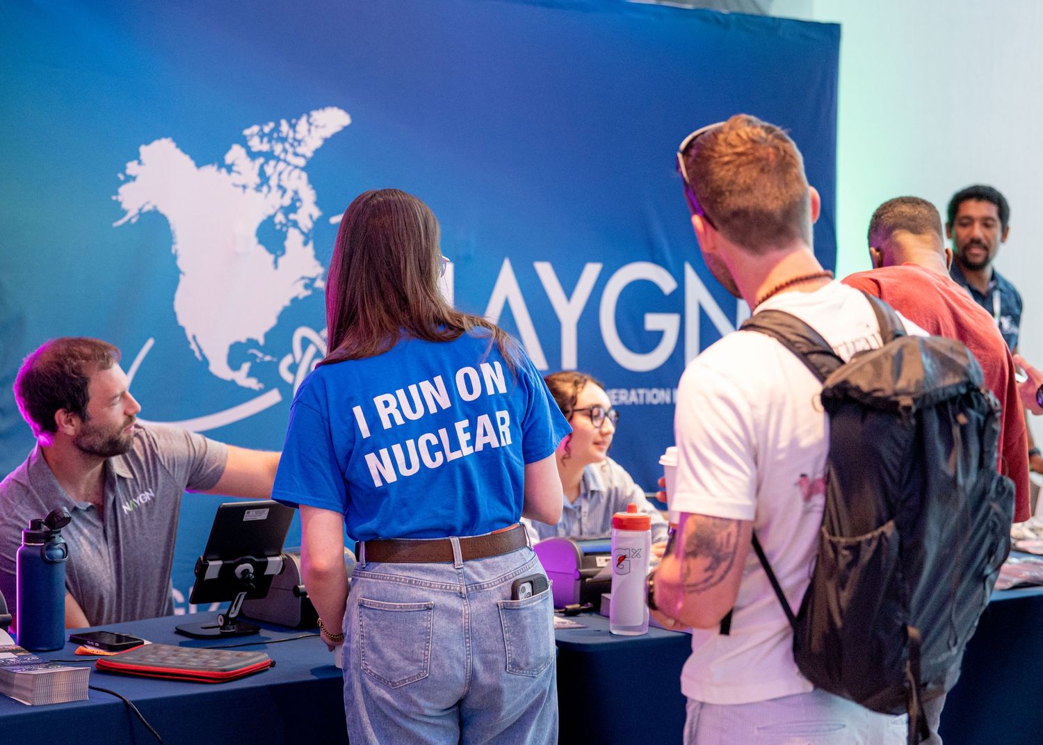 Person wearing 'I Run On Nuclear' shirt at IAYGN nuclear energy conference booth with attendees.