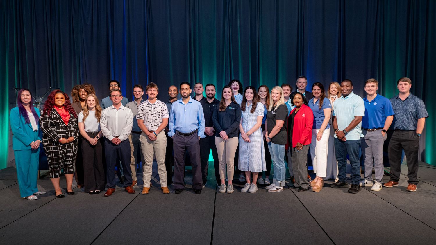 A diverse group of professionals posing together on a stage with blue curtain backdrop at a corporate event.