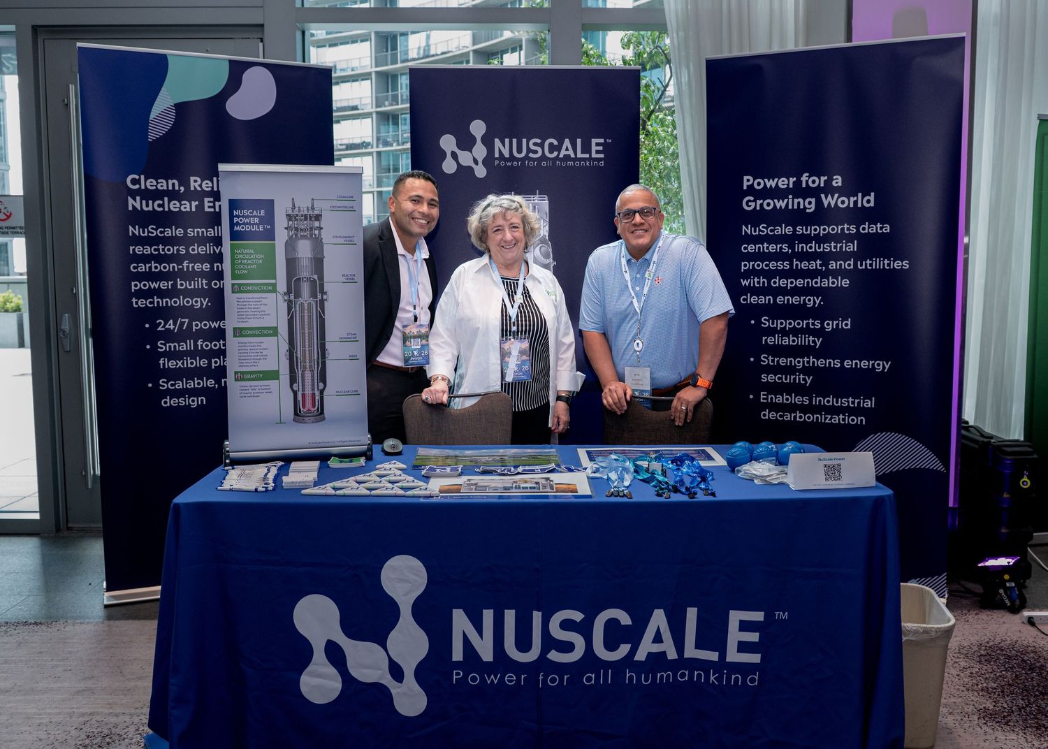 Three professionals pose at a NuScale Power trade show booth displaying nuclear energy solutions and branding materials.