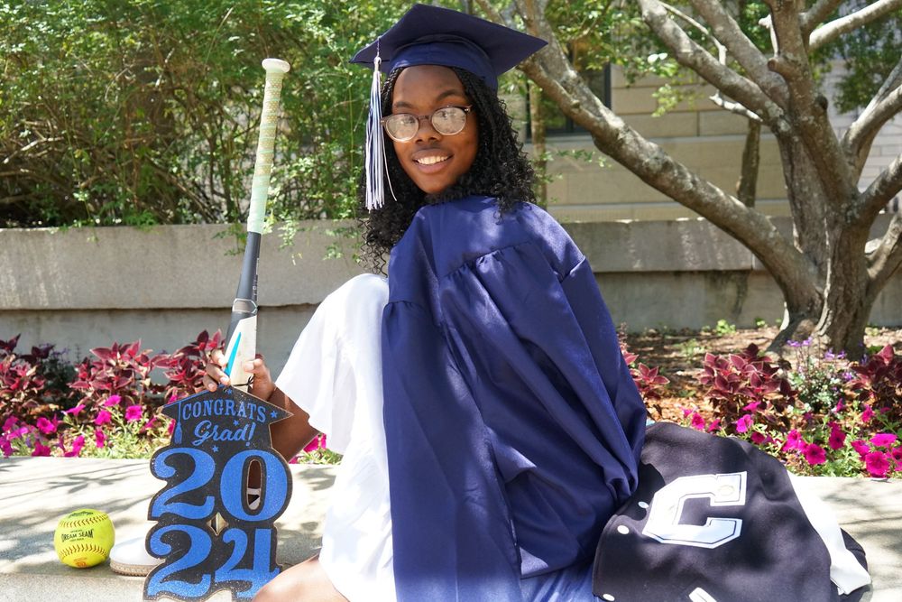 Young woman in blue graduation cap and gown poses outdoors with a Class of 2021 grad decoration.