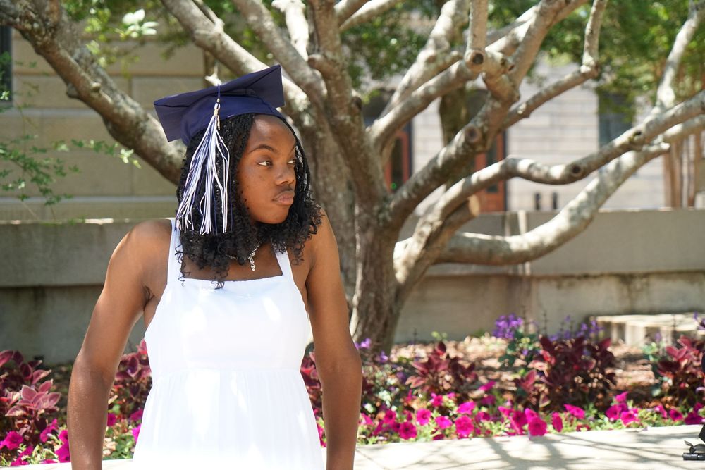 Young Black woman in white dress and navy graduation cap smiles outdoors near flowering garden and trees.