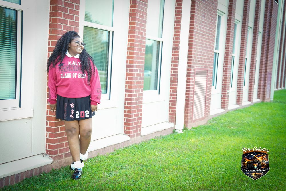 Young woman in red sweatshirt and black skirt posing outdoors beside a brick building on green grass.