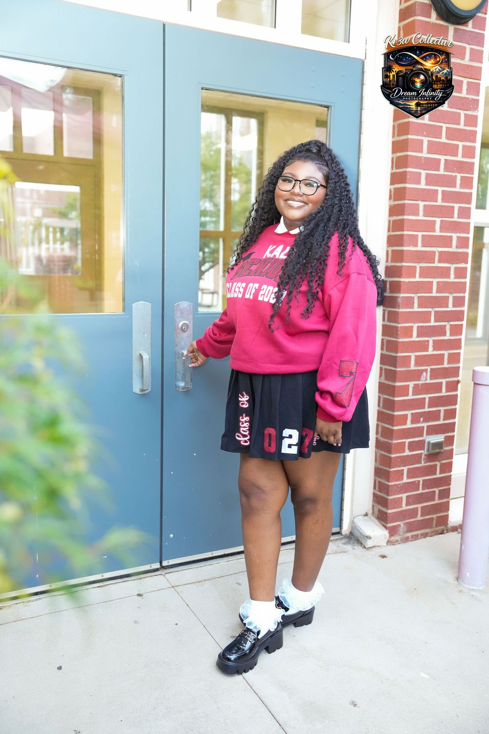 A young woman poses in a pink Class of 2027 sweatshirt, black pleated skirt, and chunky black shoes outside a brick building.