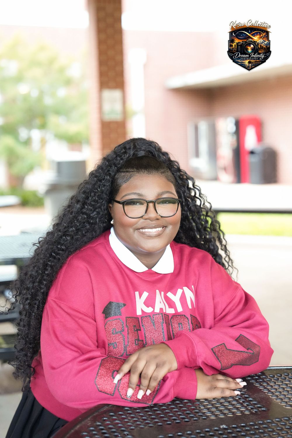Young woman in pink Kalyn sweatshirt with curly hair and glasses smiling outdoors at a table.