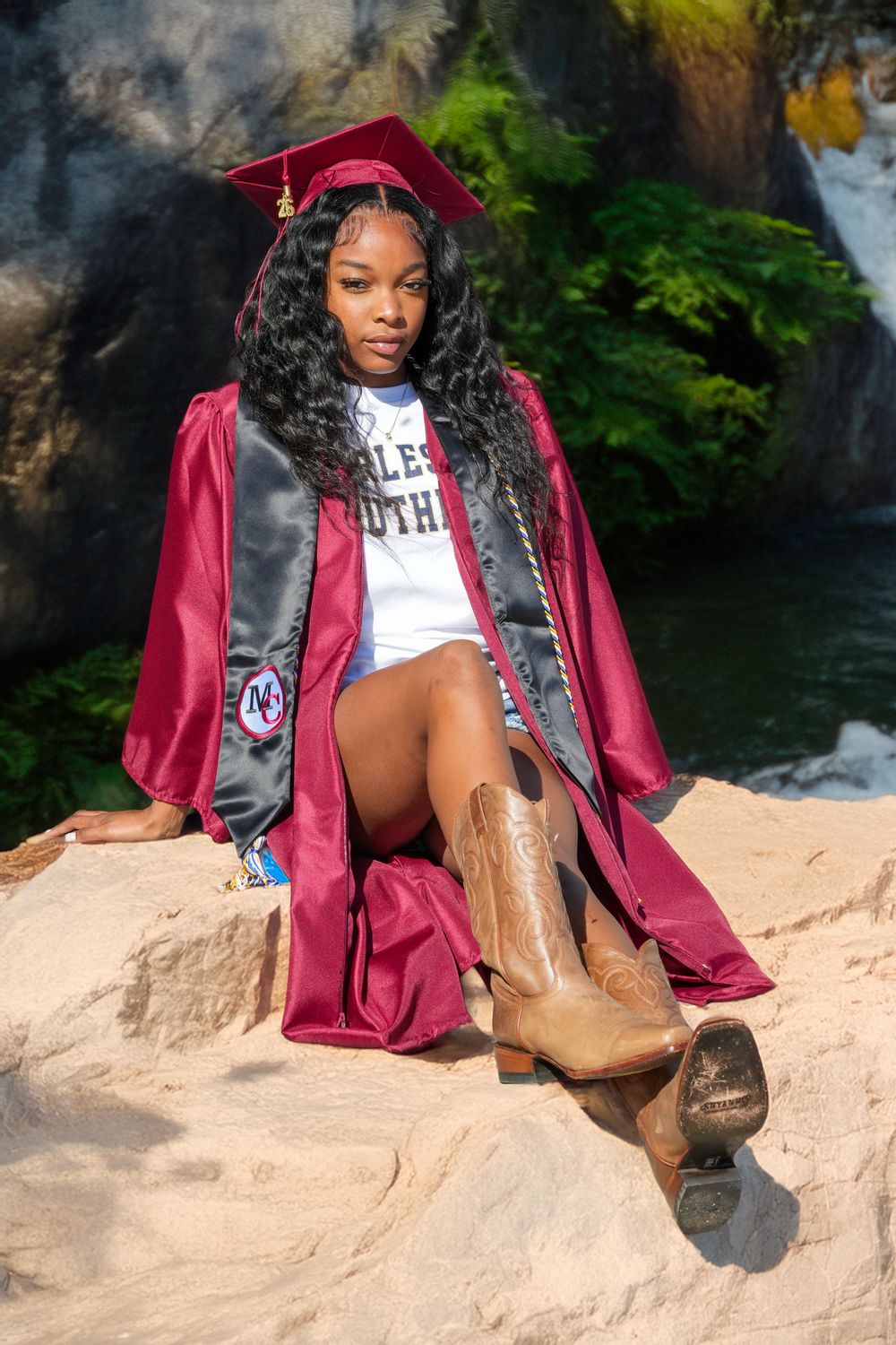 Young woman in maroon graduation cap and gown with cowboy boots sits on rocks outdoors.