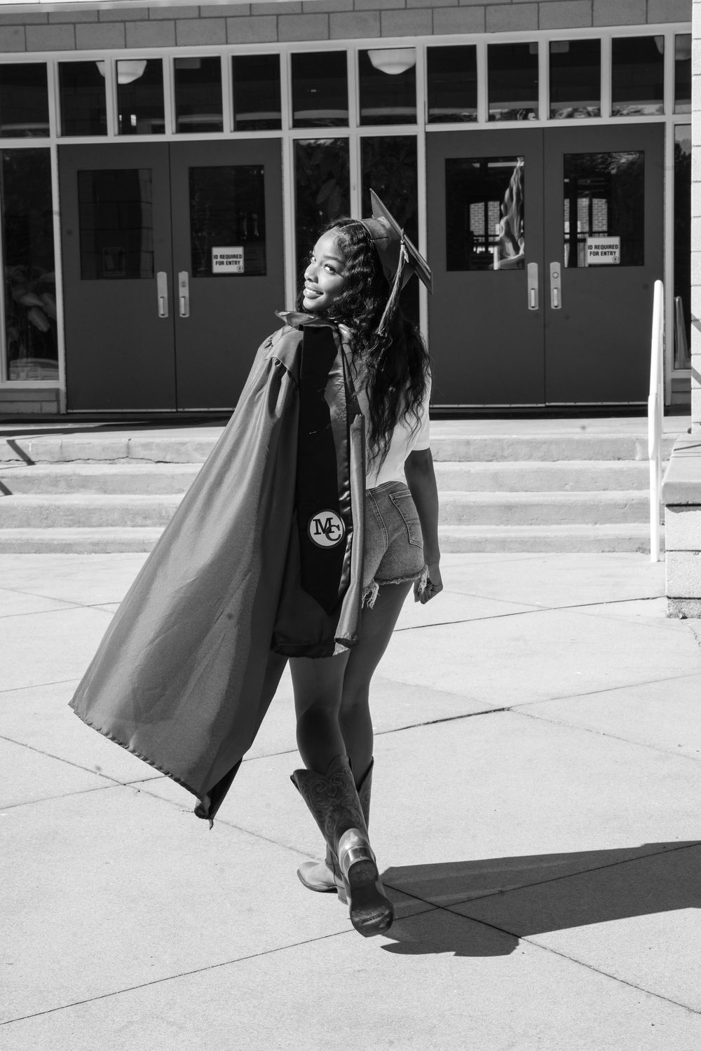Black and white photo of a person in a superhero cape and mask posing outdoors near a building entrance.