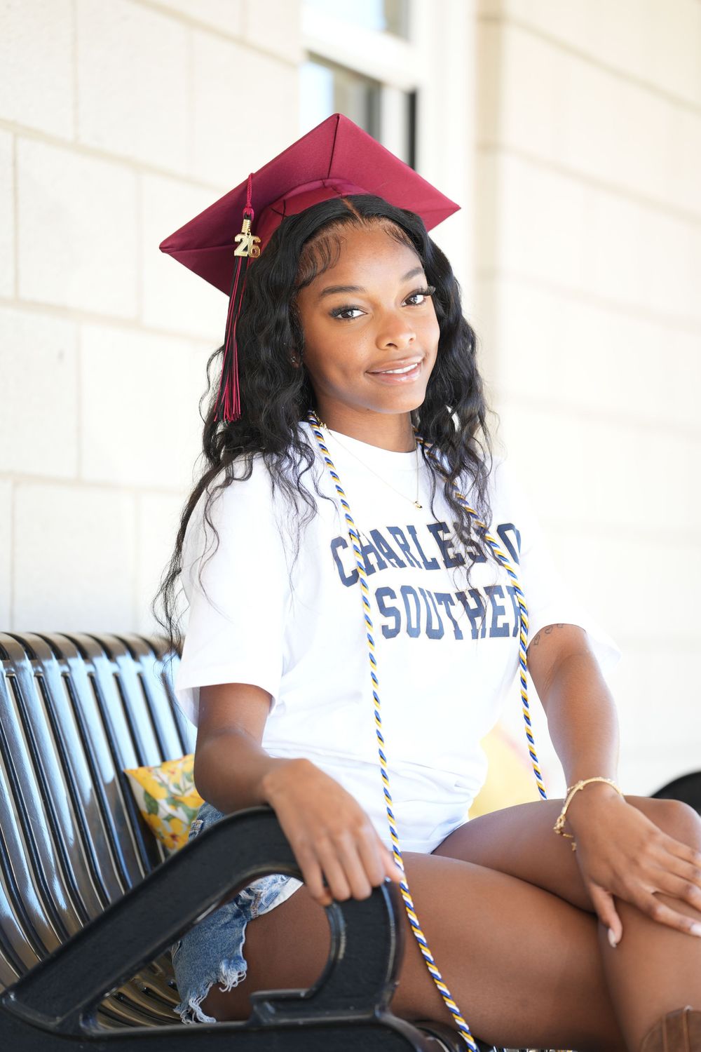 Young woman in graduation cap and Charleston Southern shirt sits on bench smiling, celebrating academic achievement.