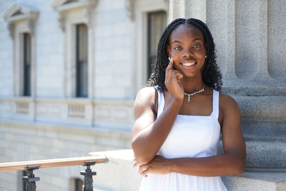 Young woman in white dress smiling confidently outdoors near a classical stone building with columns.